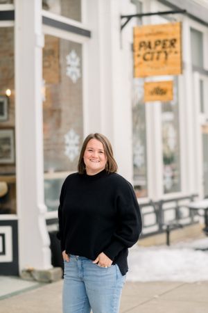 Person in black sweater standing outside Paper City storefront on sidewalk.