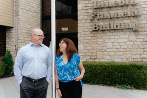 Two professionals chat while standing outside a brick office building.