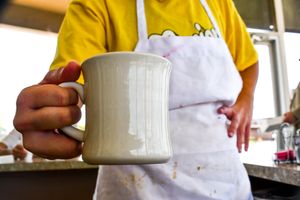 White ceramic coffee mug being held in front of white apron at a cafe.