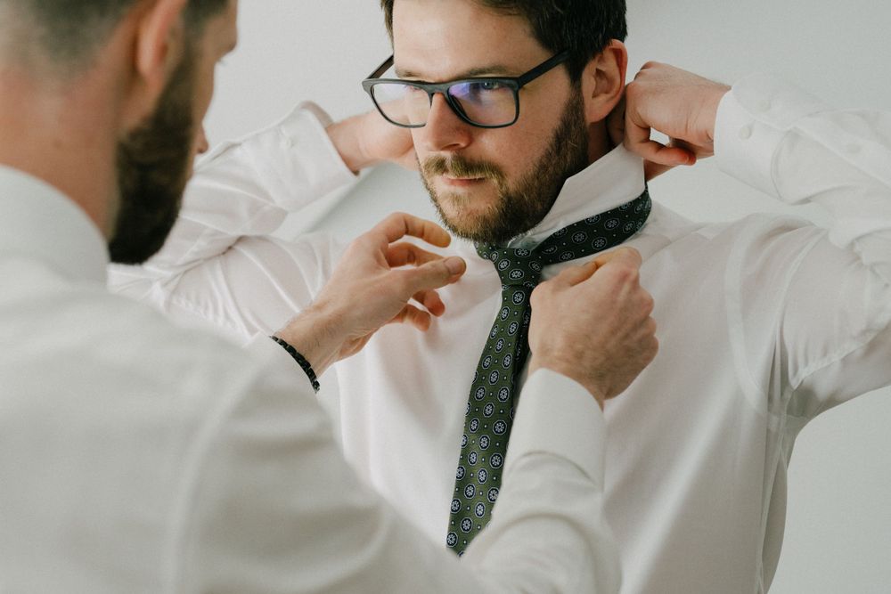 Someone helps adjust a gray tie on a person wearing glasses and a white dress shirt.