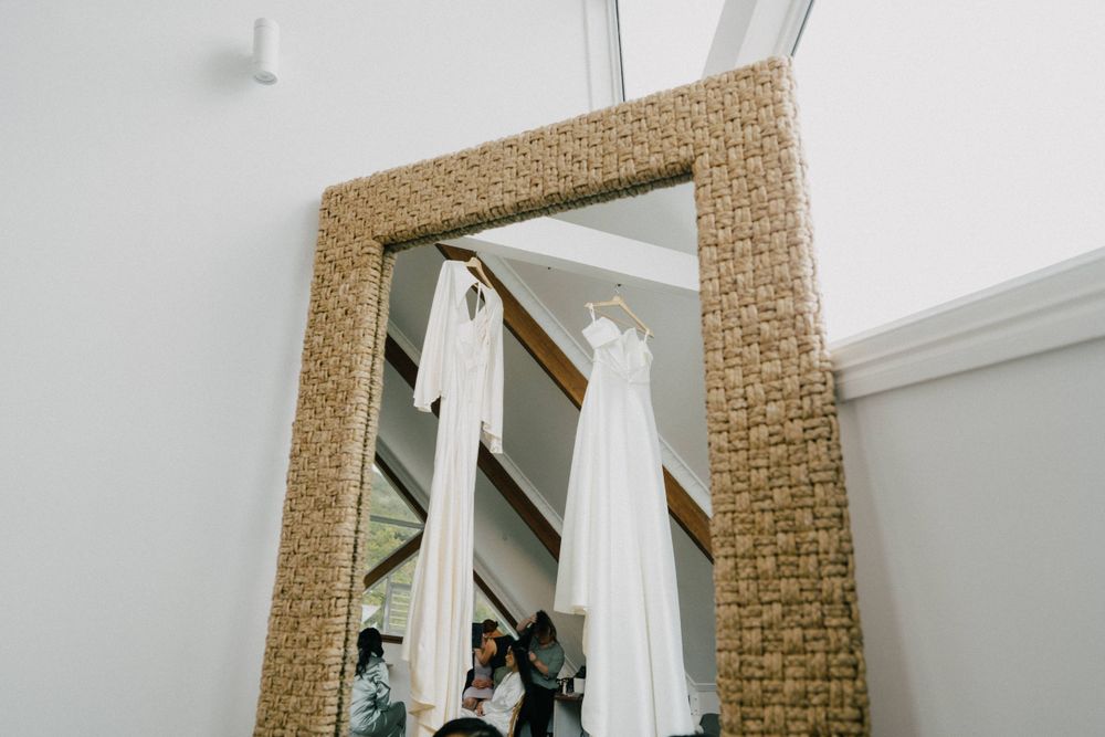 A wedding dress hangs reflected in a woven rattan-framed mirror against a white wall.