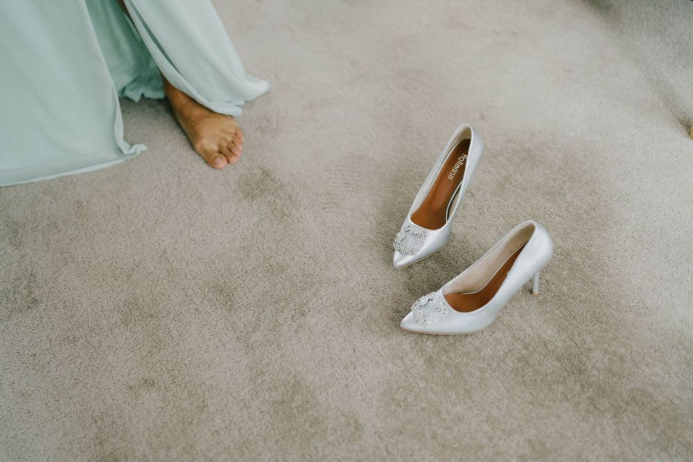 White bridal heels sit on beige carpet next to the hem of a light dress.