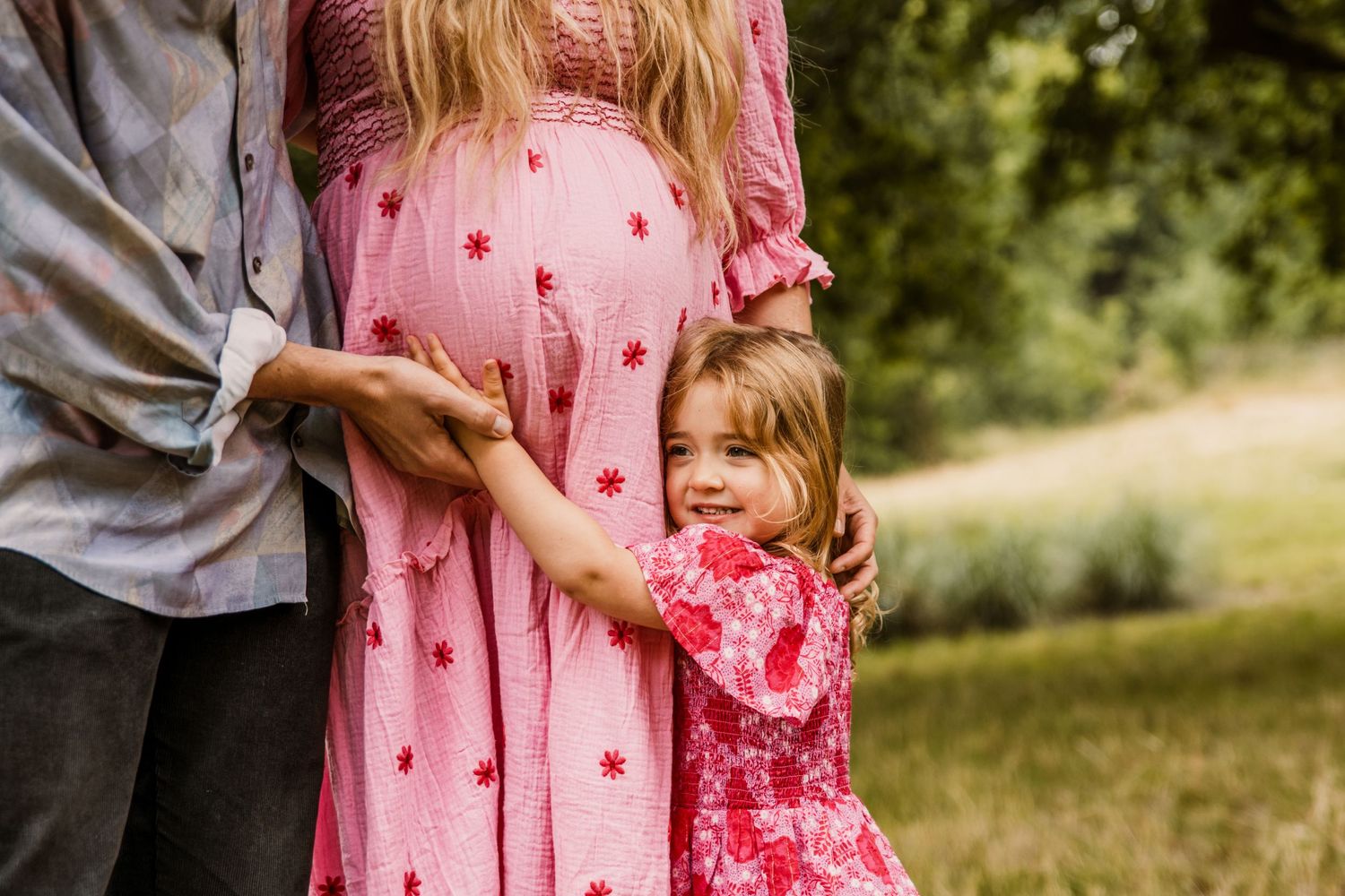 A young girl hugs her mother's baby bump in a pink dress on a summer meadow.