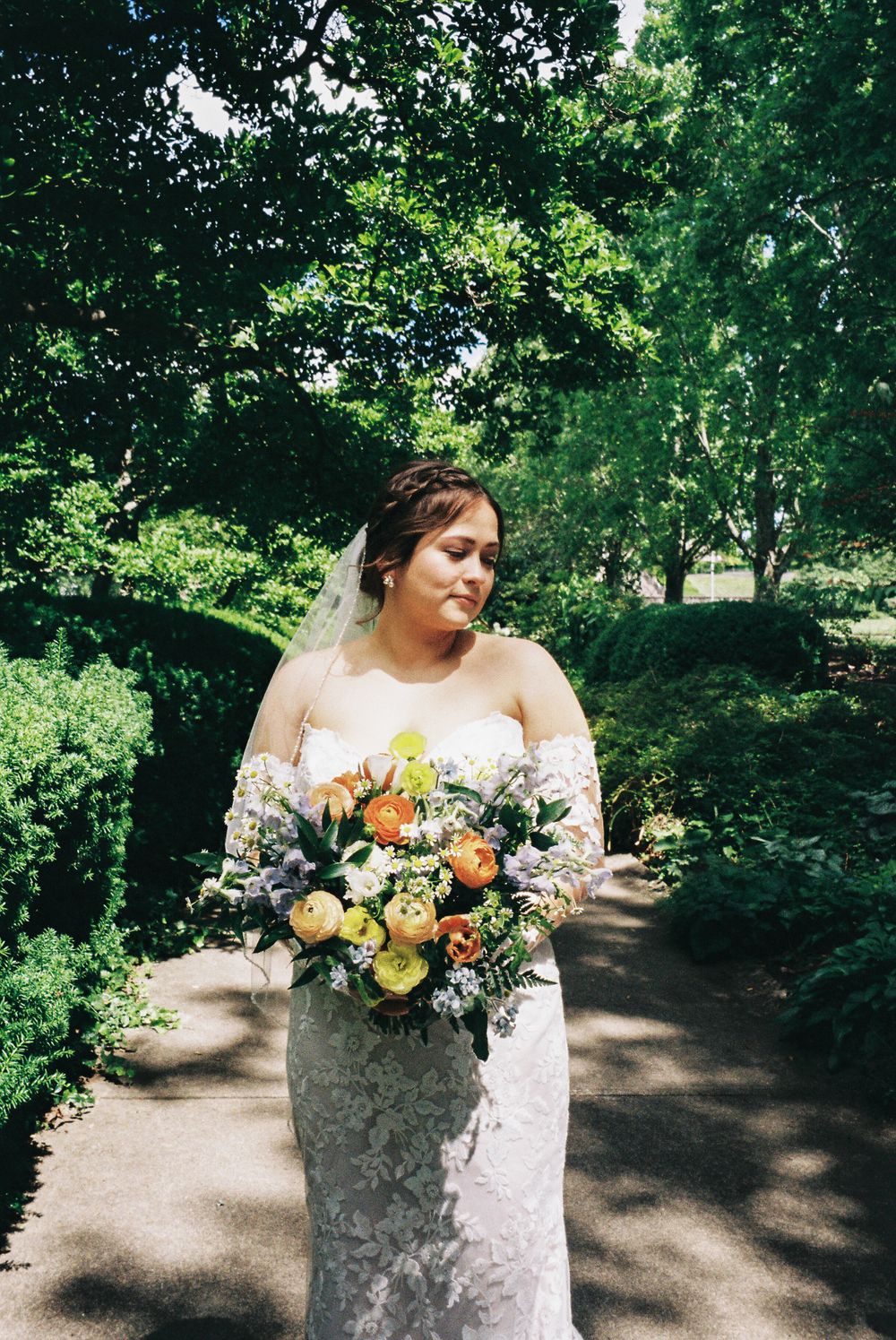 A bride in a strapless lace gown holds a vibrant yellow and purple wildflower bouquet in a garden setting.