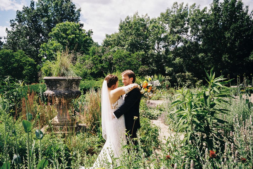A romantic wedding photo in a lush garden with decorative stone fountain in the background.