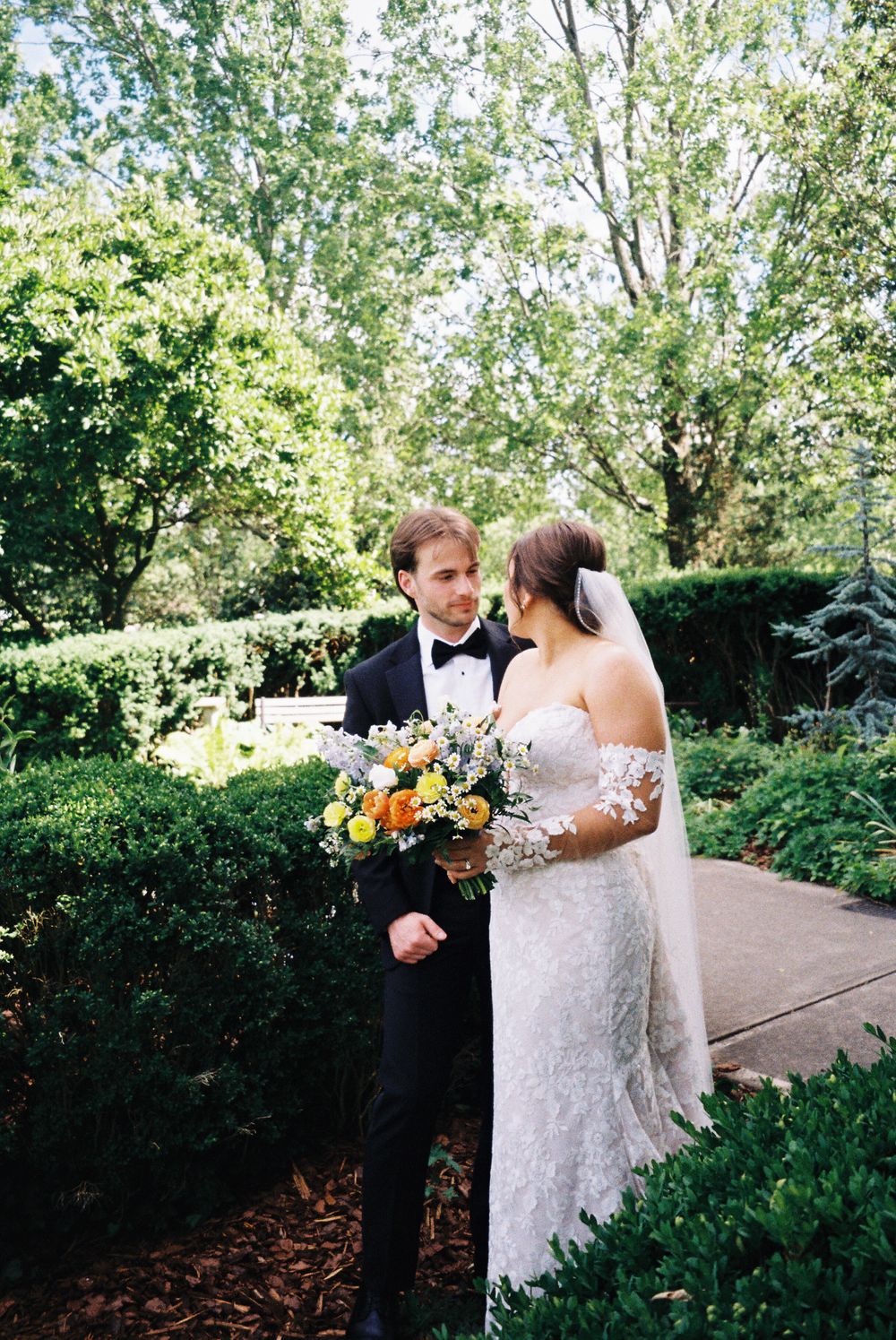 A wedding couple poses in a manicured garden setting with mixed floral bouquet.