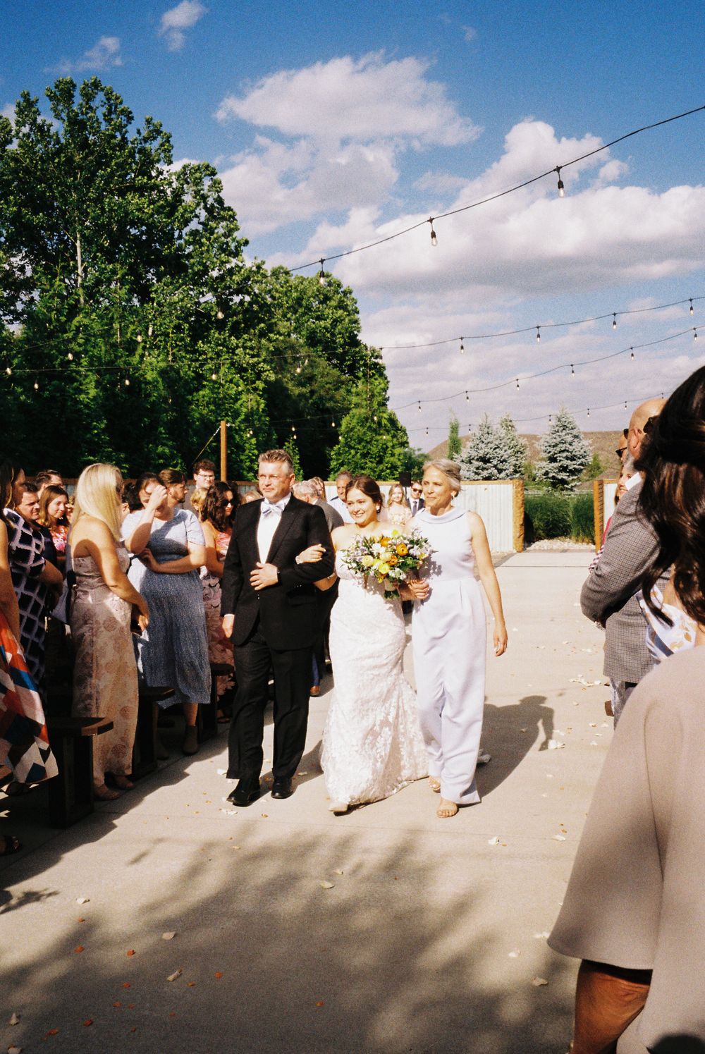 Wedding procession walking outdoors with string lights overhead on a sunny day.