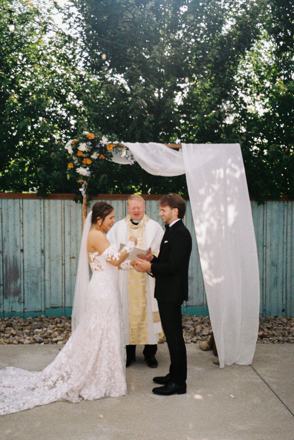 A wedding ceremony under a white fabric arch with floral decorations against a blue fence backdrop.