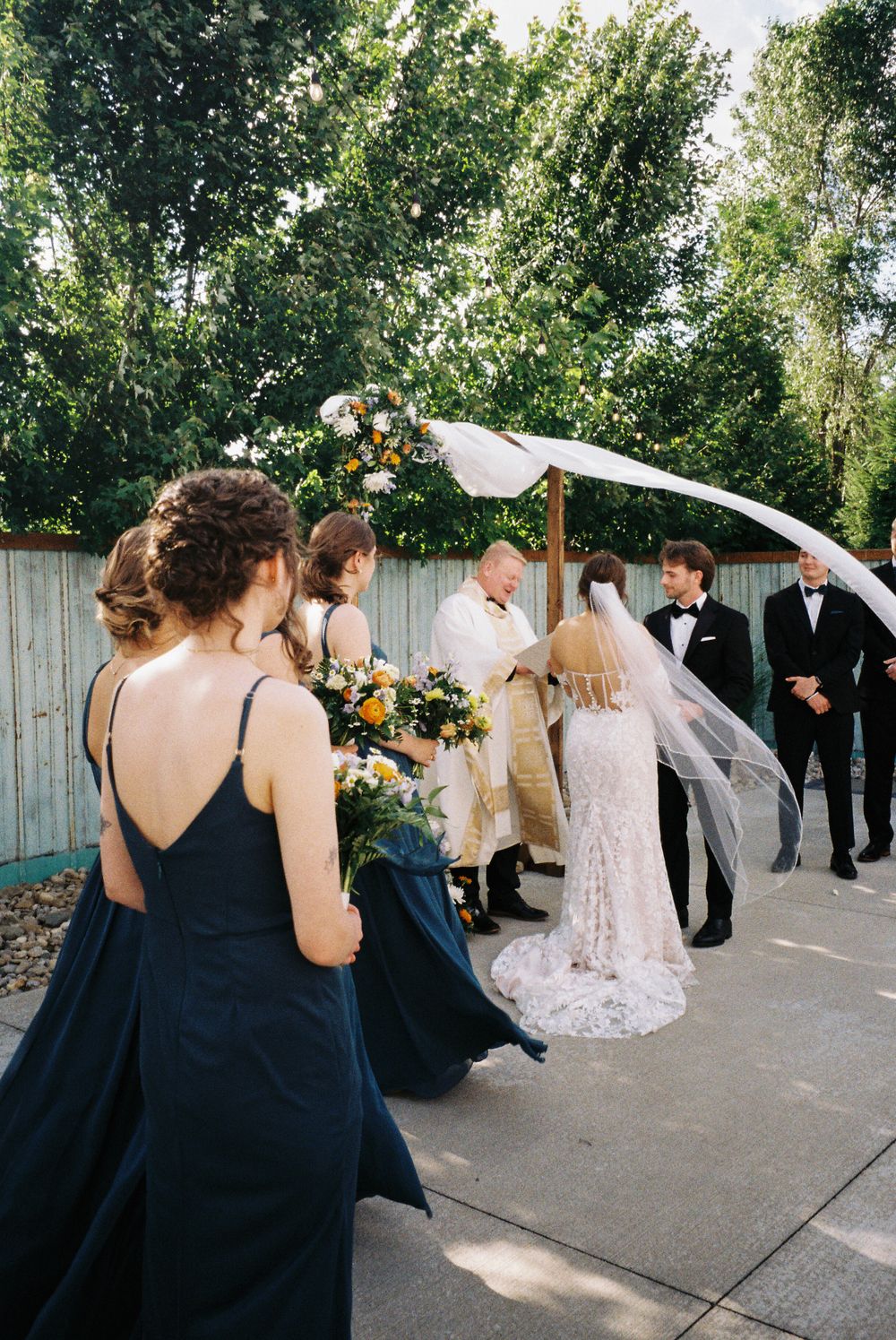 An outdoor wedding ceremony with bridesmaids in navy dresses and a white ceremony arch adorned with flowers.