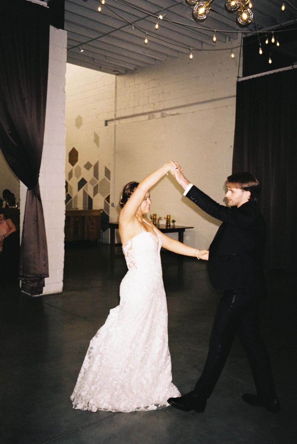 A couple dancing in an industrial venue with string lights overhead during their wedding reception.