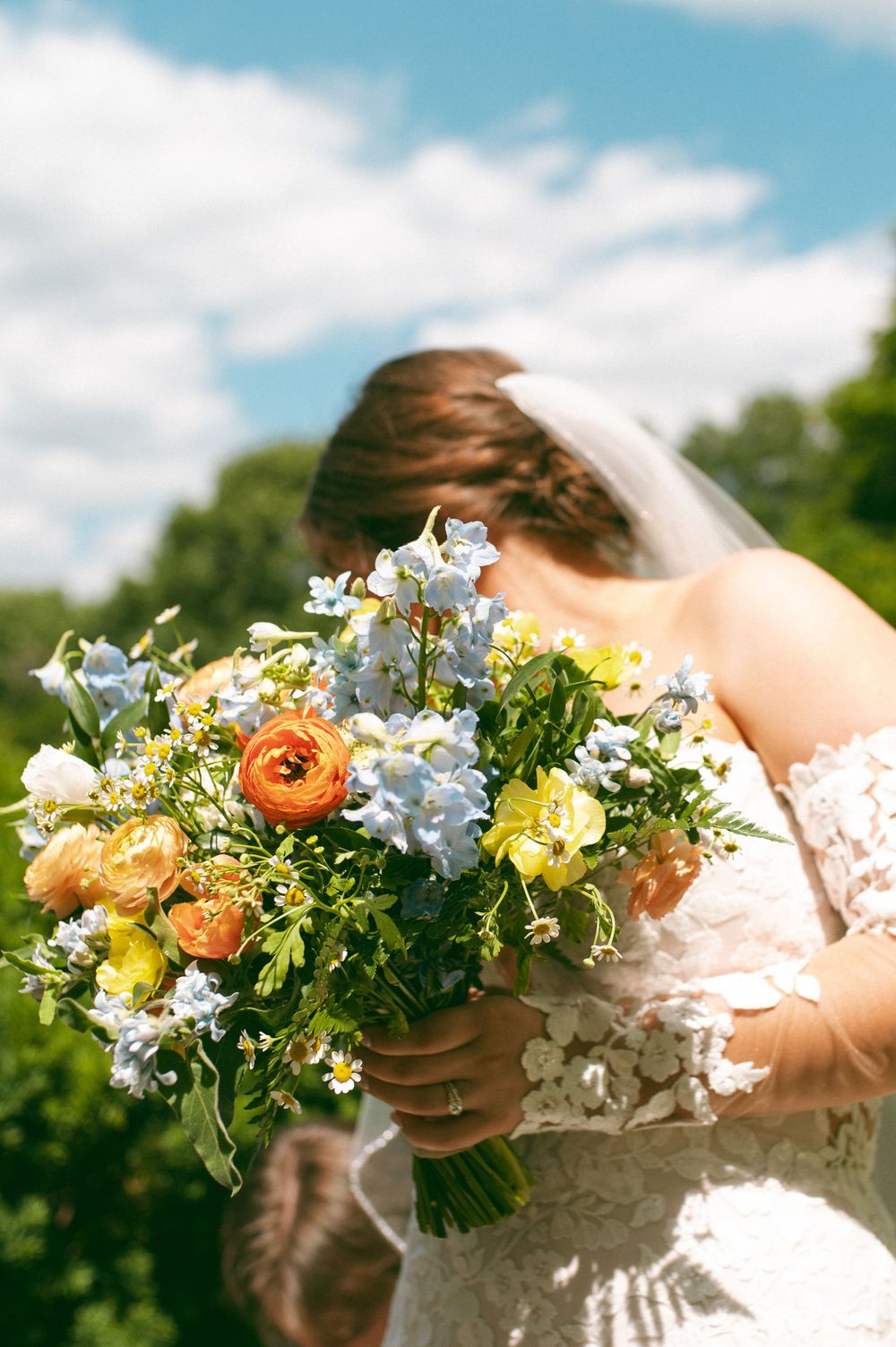 Vibrant bridal bouquet with orange ranunculus and blue delphinium against bright summer sky.