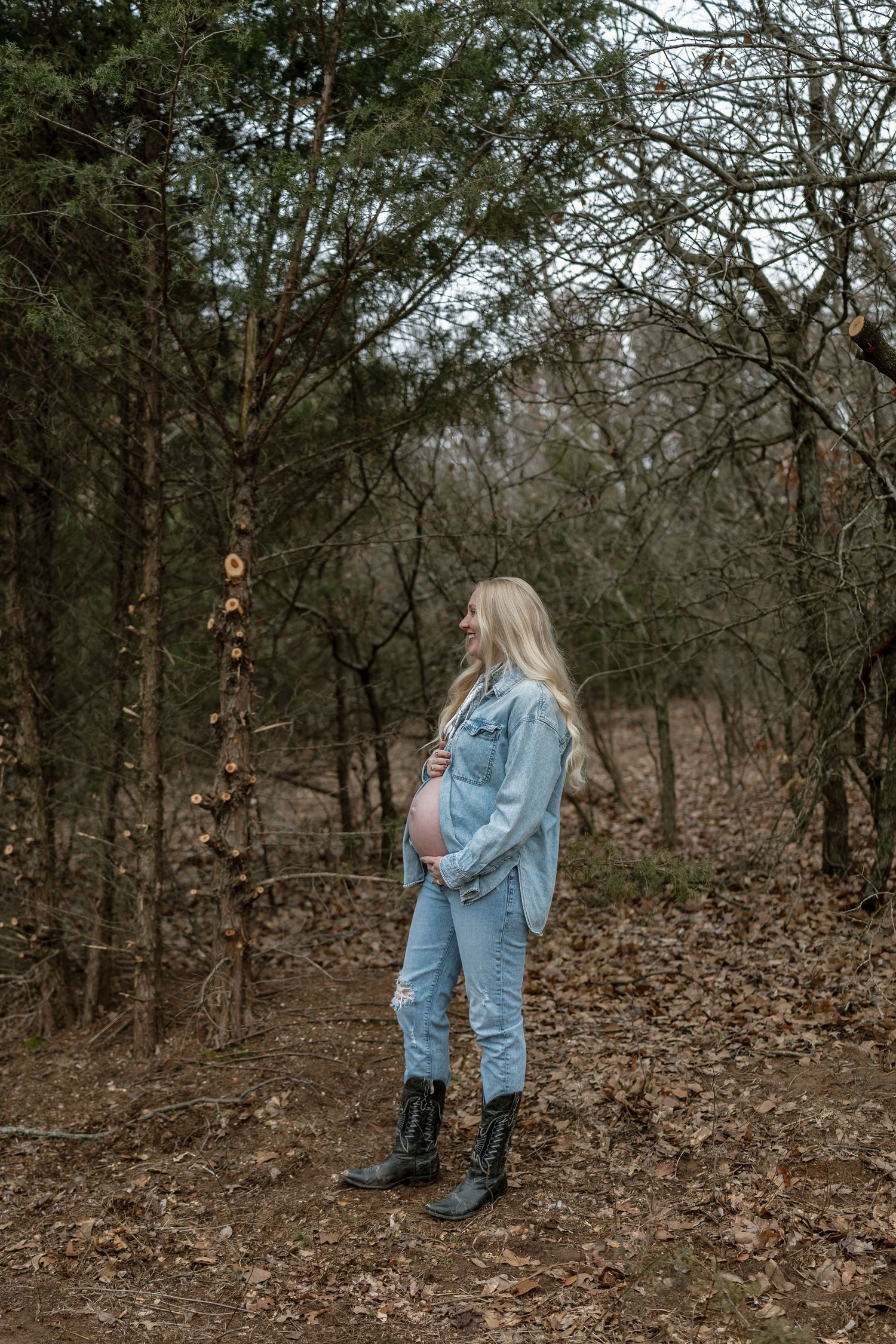 Expectant mother in denim jacket and jeans poses in a wooded forest setting during winter.