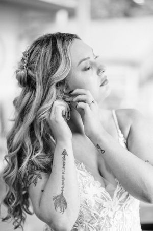 Black and white portrait of a bride in lace dress with flowing curled hair and visible tattoos putting her earrings in