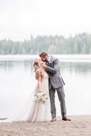 A romantic wedding couple shares intimate moments by a serene lakeside with evergreen trees in the background.