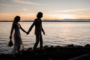 Silhouettes of people walking along a rocky beach at sunset with mountains and ocean in the background.