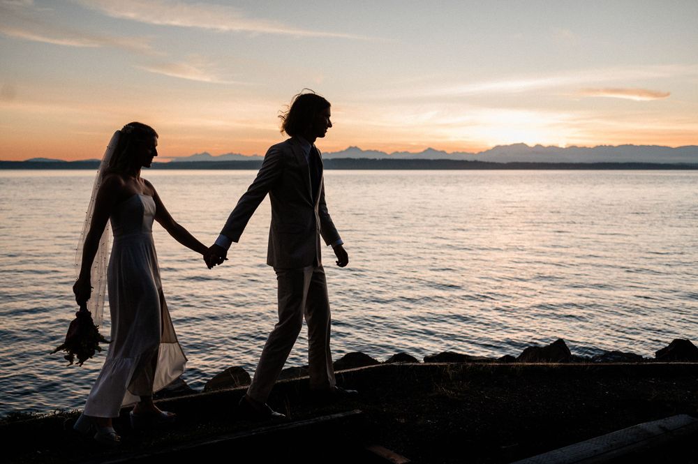 Silhouettes of people walking along a rocky beach at sunset with mountains and ocean in the background.