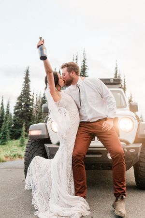 A couple in wedding attire kisses while sitting on a jeep with evergreen trees in the background. Photo by Pine + Vow