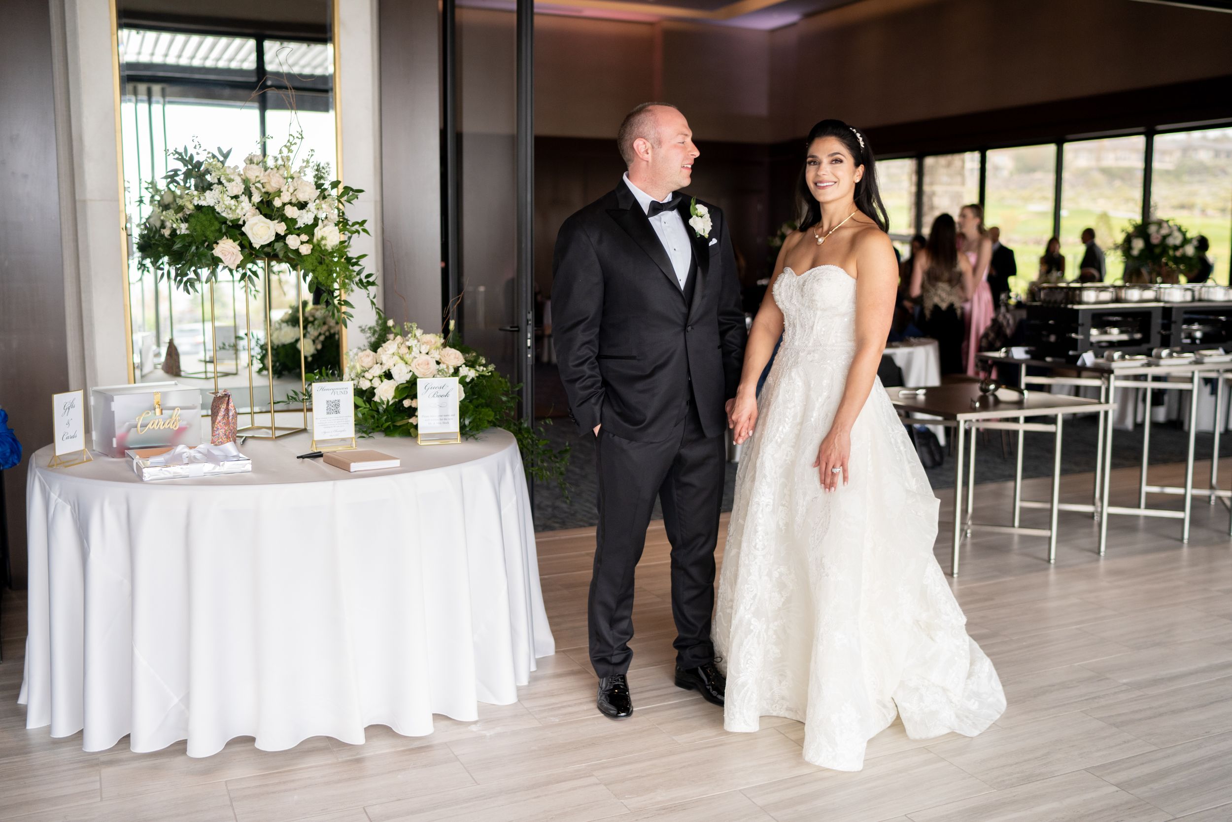 A couple in formal wedding attire stand together in an elegant reception venue with white table decor and floral arrangements
