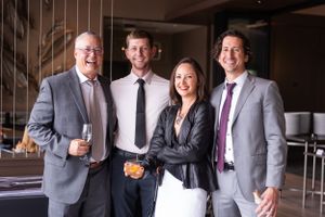 Four professionally dressed colleagues stand together smiling at a wedding with drinks in hand.