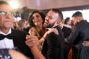 Happy couple laughing and dancing together at a formal event with other guests visible in the background.