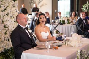 Elegant couple sits at head table during wedding reception with white floral arrangements and formal decor.