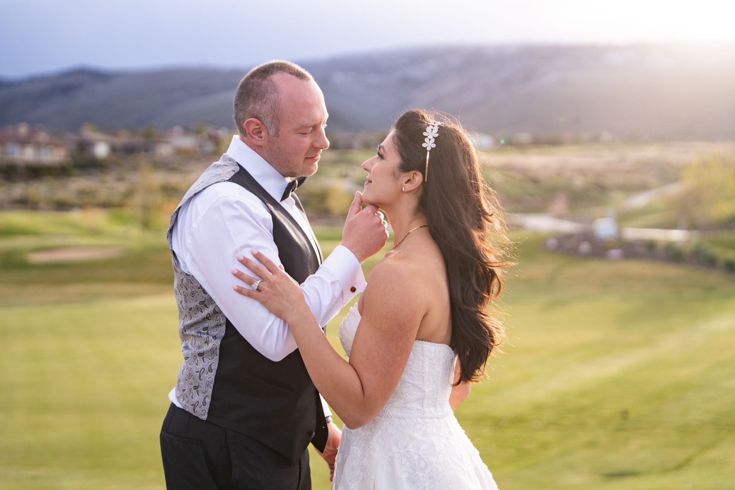 A couple shares a romantic moment on a golf course with mountains and golden sunlight in the background at The Club at Arrowcreek