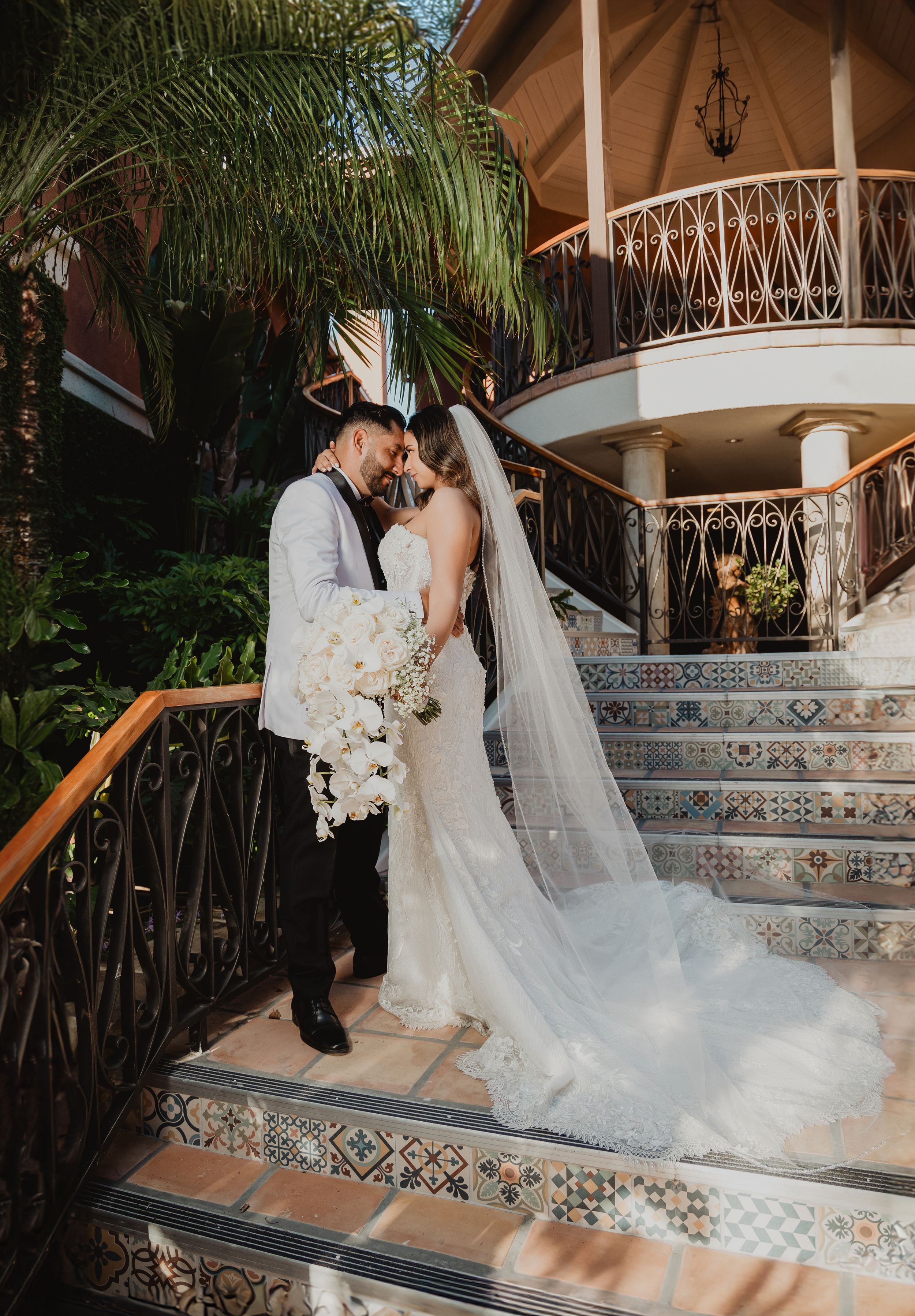 Wedding couple standing on ornate tiled stairs with palm trees and Spanish-style architecture.