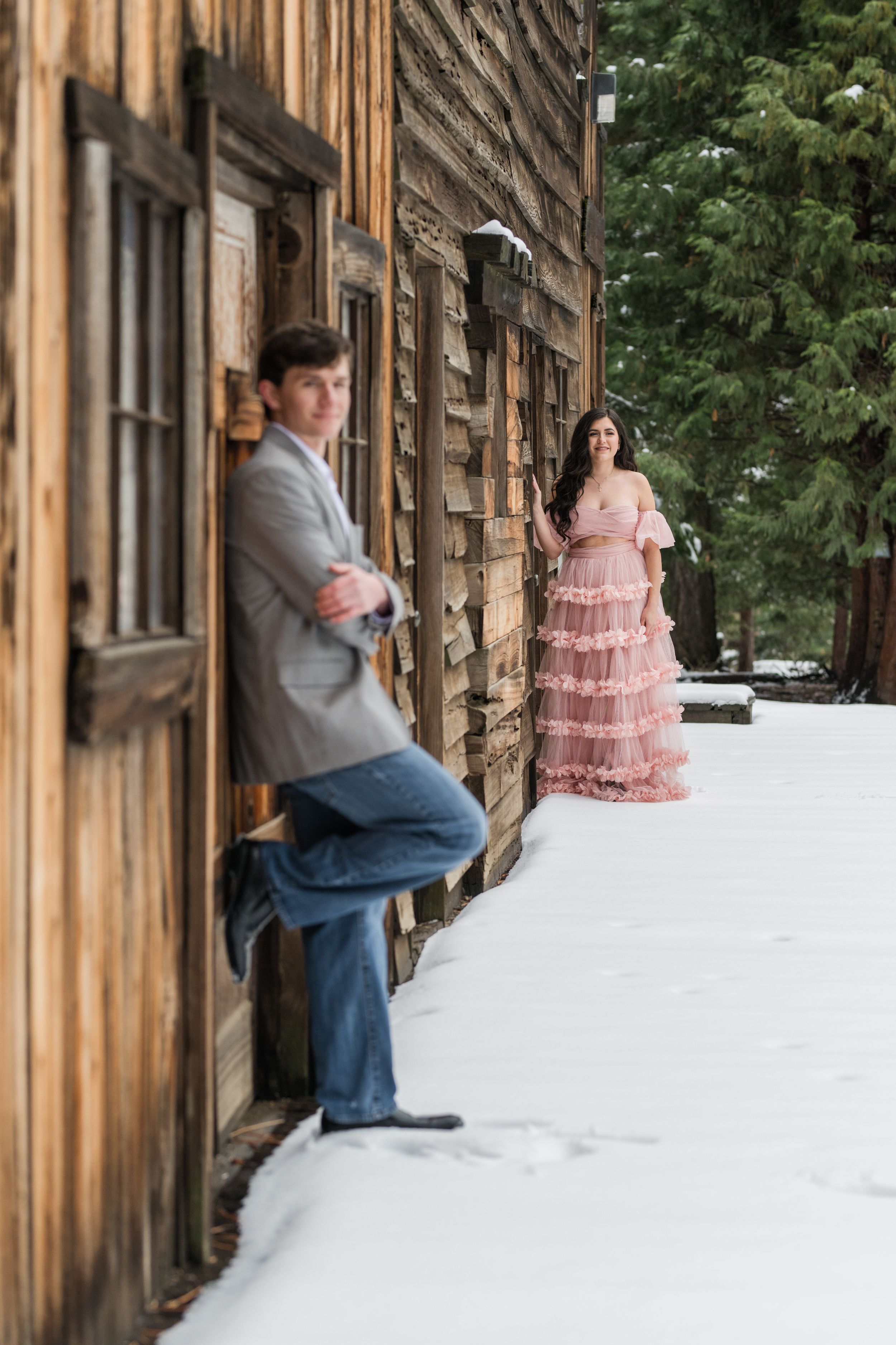 Stylish couple posing by rustic wooden cabin in winter with snow on the ground.