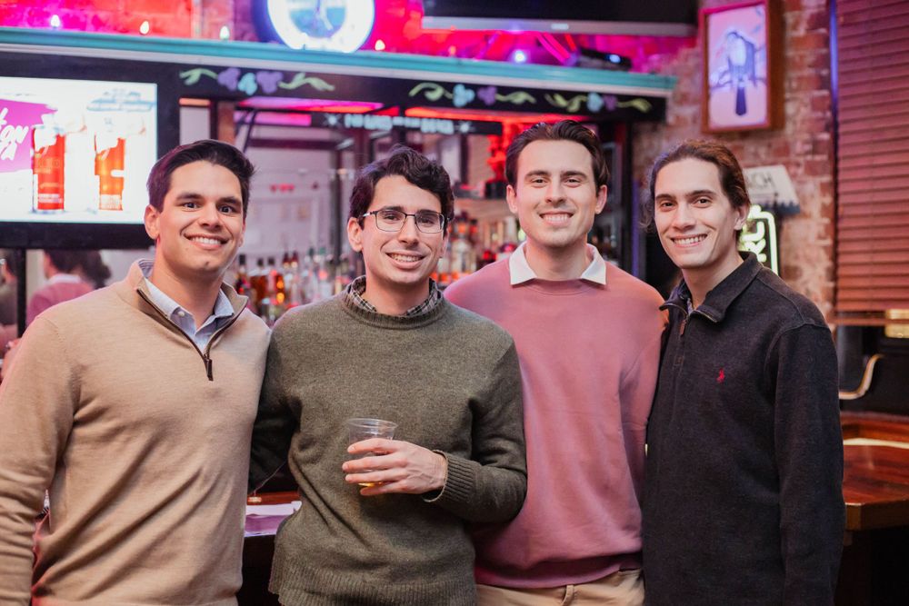 Four friends in casual sweaters smiling while gathered together at a bar with neon lighting in the background.