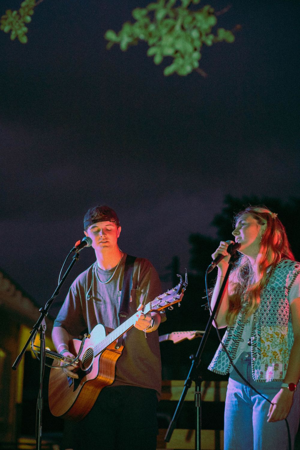 Musicians performing on an outdoor stage at night with acoustic guitar and microphones.