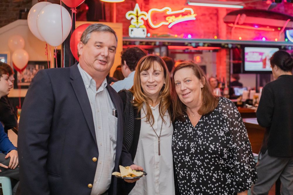 Three people smile together at a casual party with red neon lighting and balloons in the background.