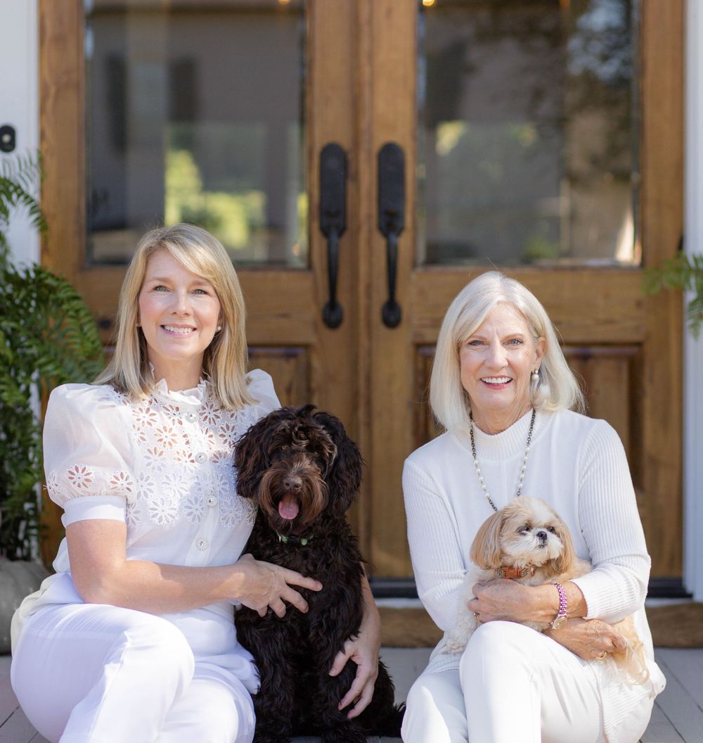 Two people in white outfits sit outdoors on chairs with their pets, a dark-colored dog and small brown dog.