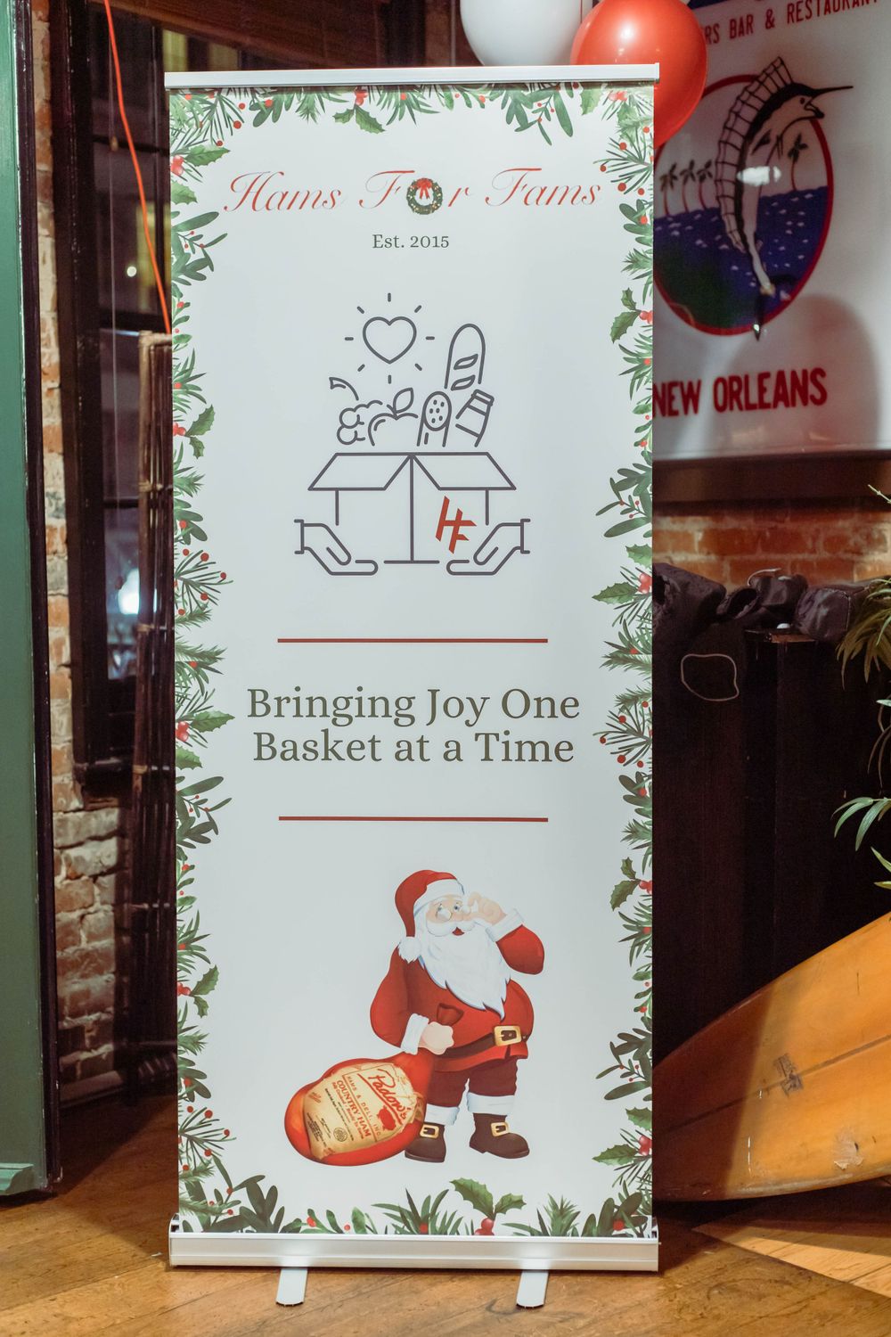 A white banner stand displays 'Hams For Fams' charitable message with holly border and Santa Claus illustration delivering baskets.