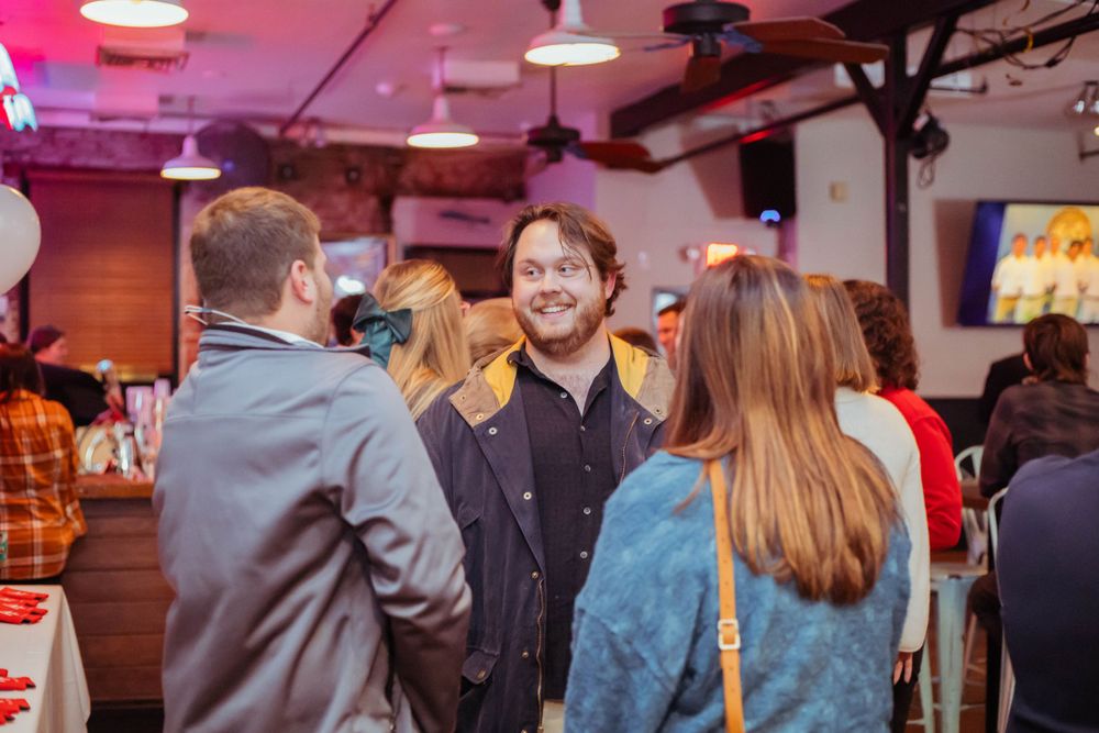 People socializing at a casual event in a venue with pink lighting and industrial ceiling.