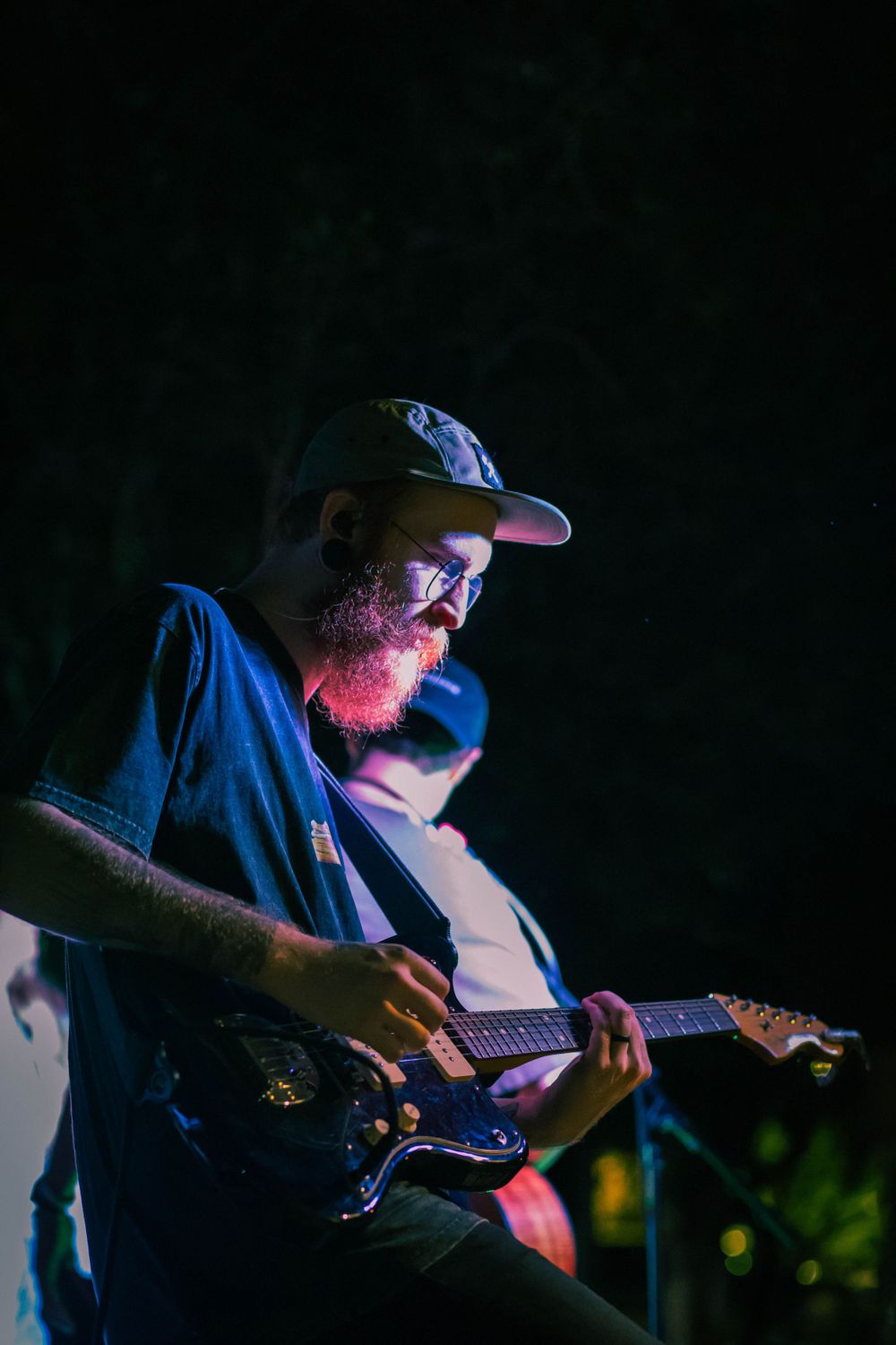 Guitar player with beard and baseball cap performing under dramatic blue lighting.