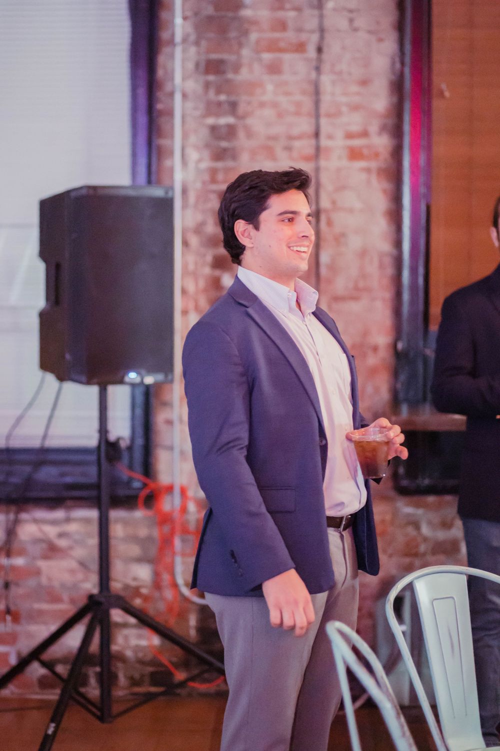 A person in a navy blazer and light shirt stands smiling with a speaker in the background at an indoor venue.