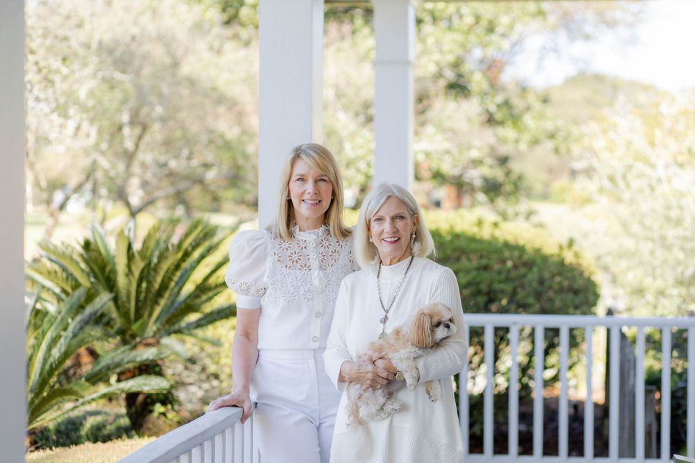 Two people pose together on a white porch with tropical foliage and greenery in the background.
