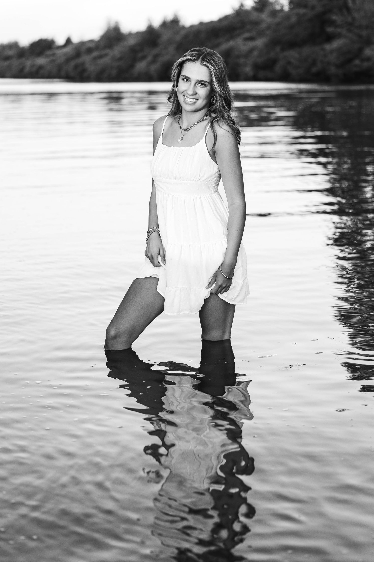 Smiling young woman in white dress stands in shallow lake water wearing boots in black and white portrait.