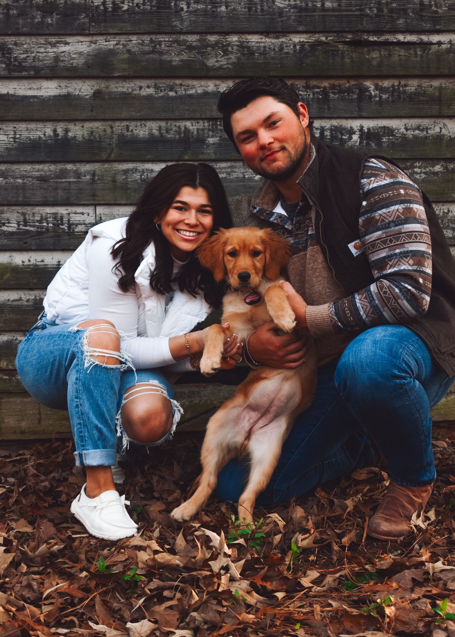 A couple sits with their golden retriever puppy against a rustic wooden wall surrounded by fallen leaves.