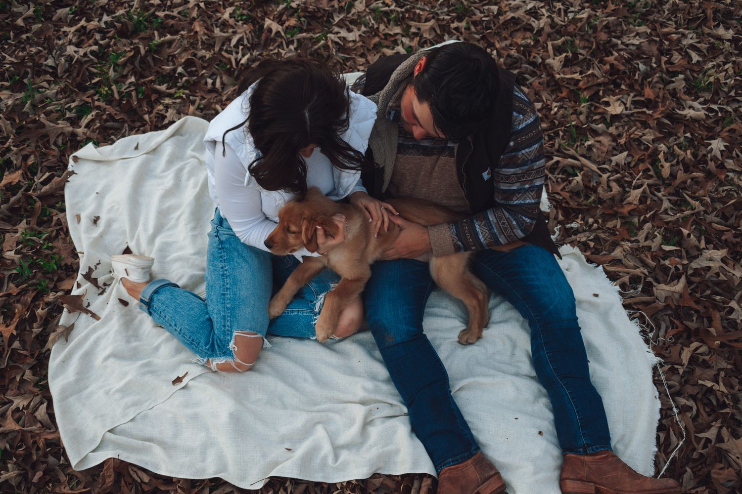 A couple relaxes on a white blanket with their puppy among scattered fall leaves.
