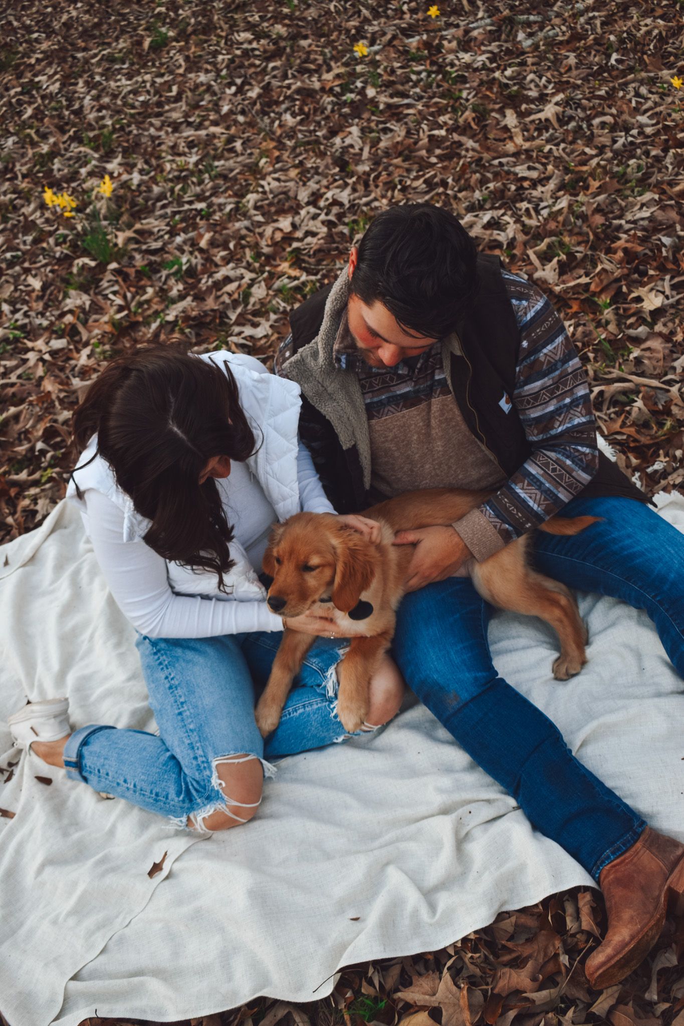 A couple plays with their puppy while sitting on a white blanket covered in autumn leaves.