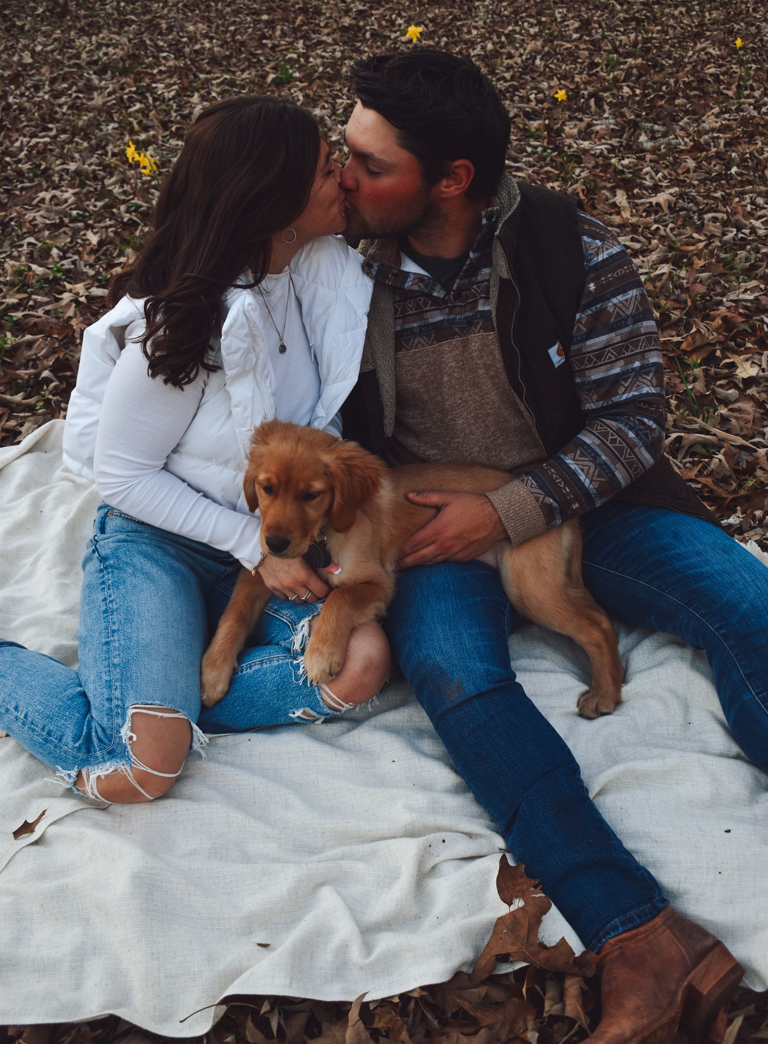 A romantic couple shares a moment with their golden puppy on a blanket surrounded by autumn leaves.