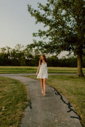 Young woman in white sundress walking along curved path in green park during golden hour sunset.