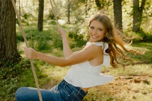 Smiling blonde girl in white top and jeans swinging on rope swing in sunlit green forest.