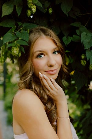 Young woman with green eyes and wavy hair poses gracefully among lush green hop vines outdoors.