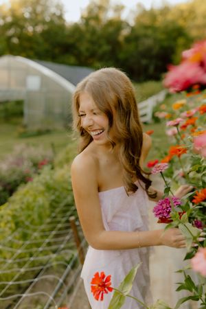 Laughing young woman in white strapless dress stands among colorful zinnia flowers in garden.