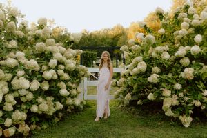 Young woman in pink dress standing among blooming white hydrangea bushes in golden evening light.