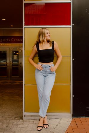 Blonde woman in black crop top and jeans leaning against colorful wall outside Performing Arts Center.
