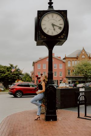Blonde woman leans against vintage town square clock post holding red roses in heels.