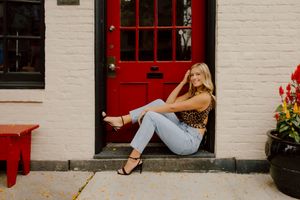 Blonde woman in leopard print top and jeans poses by red door on brick building.